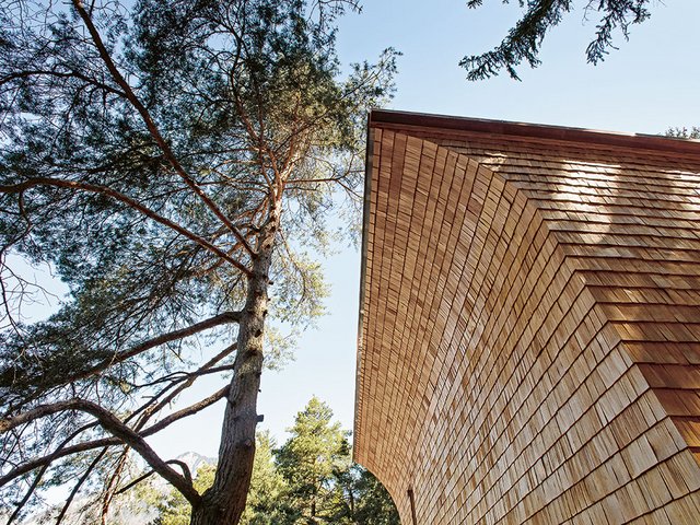 Die mit handgefertigten Lärchenschindeln geschuppte Frontfassade mit dem oben bogenartig auskragenden Bug. Im kleinen Fenster spiegelt sich die Bergwelt.