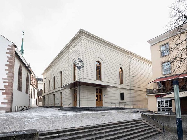 Das geschlossene Gemäuer verbirgt den Musiksaal, hinter den Rundbogenfenster liegt ein neues Foyer.