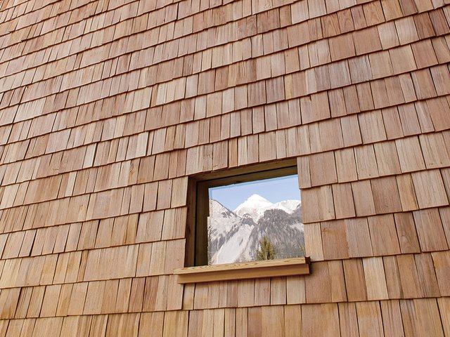Die mit handgefertigten Lärchenschindeln geschuppte Frontfassade mit dem oben bogenartig auskragenden Bug. Im kleinen Fenster spiegelt sich die Bergwelt.